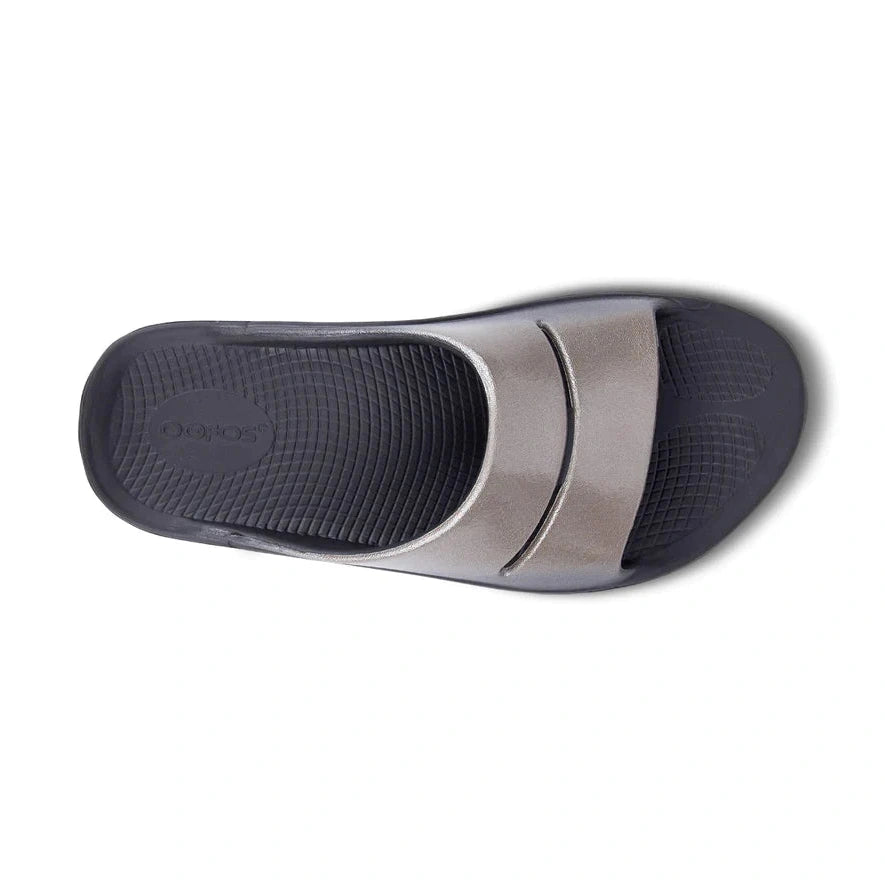 A single OOfos OOahh Luxe slide sandal shown from a top-down view, featuring a metallic silver strap and a dark gray textured footbed.