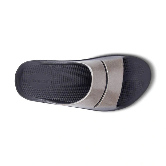 A single OOfos OOahh Luxe slide sandal shown from a top-down view, featuring a metallic silver strap and a dark gray textured footbed.