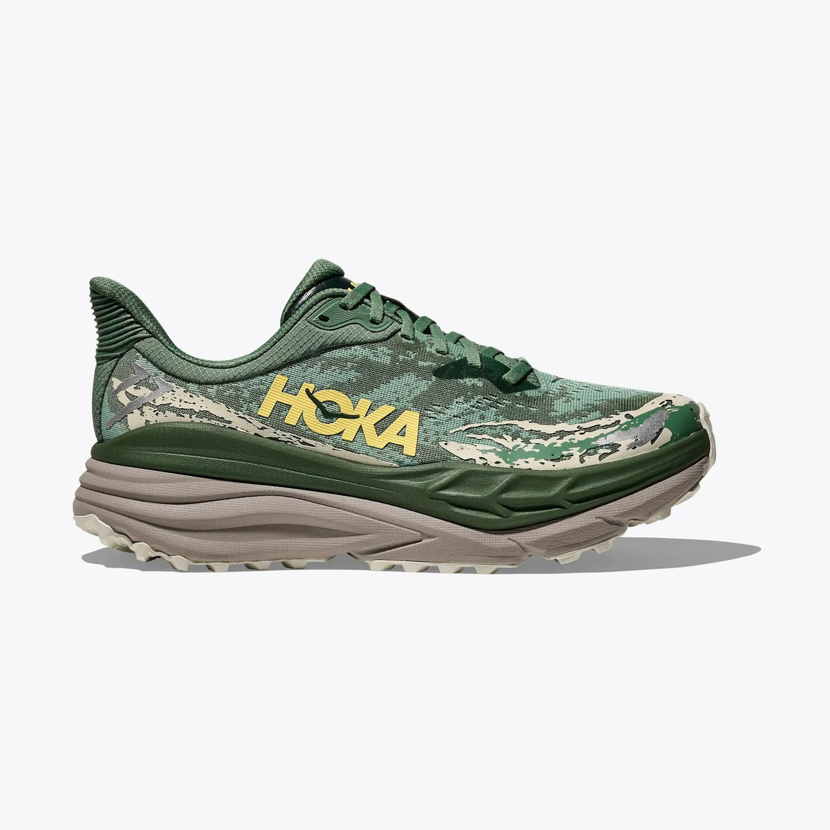 A Hoka Stinson 7 – Men's trail running shoe in green and beige camouflage with a prominent yellow HOKA logo on the side, shown against a light gray background.
