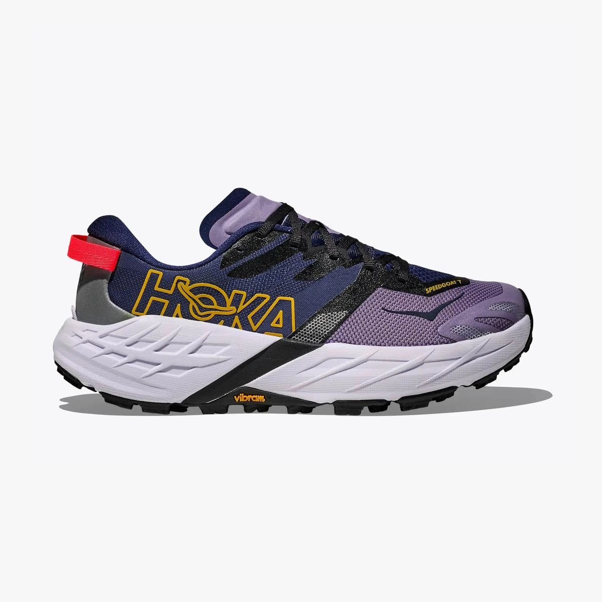 A Hoka Speedgoat 7 – Women's trail running shoe in navy blue, purple, and white, featuring a prominent yellow 'HOKA' logo on the side, a red heel pull tab, and Vibram branding on the sole.