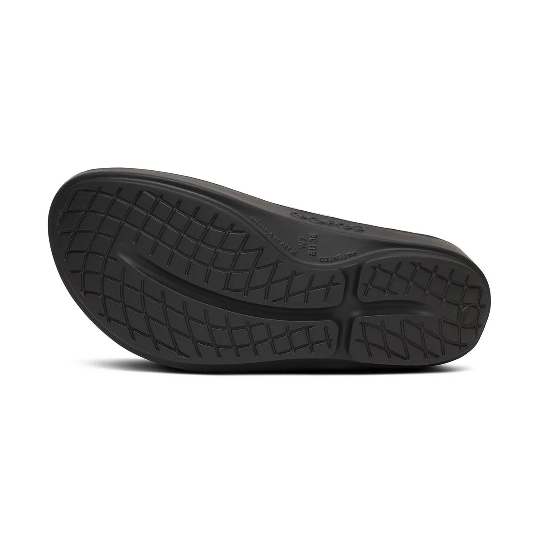 The black rubber outsole of a shoe, showing a textured tread pattern and embossed branding, photographed from a direct overhead angle on a white background.