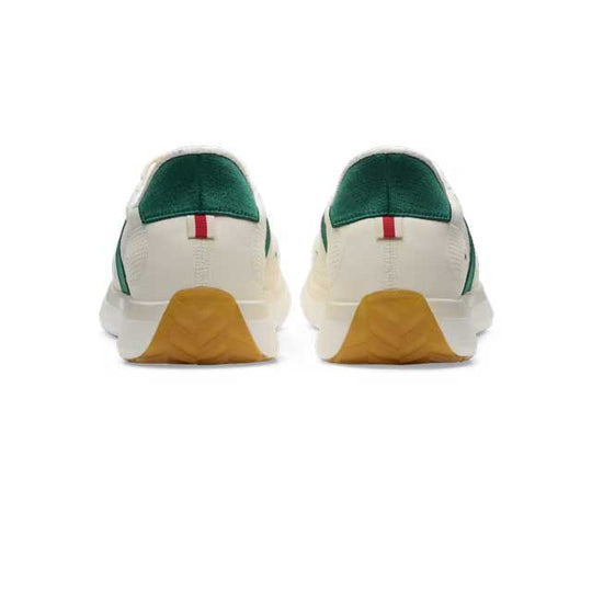 A pair of Tracksmith Eliot Ryder – All Gender low-top sneakers shown from the back, featuring an off-white leather upper, a dark green suede heel tab, a small red and green leather detail, and a textured tan rubber outsole.