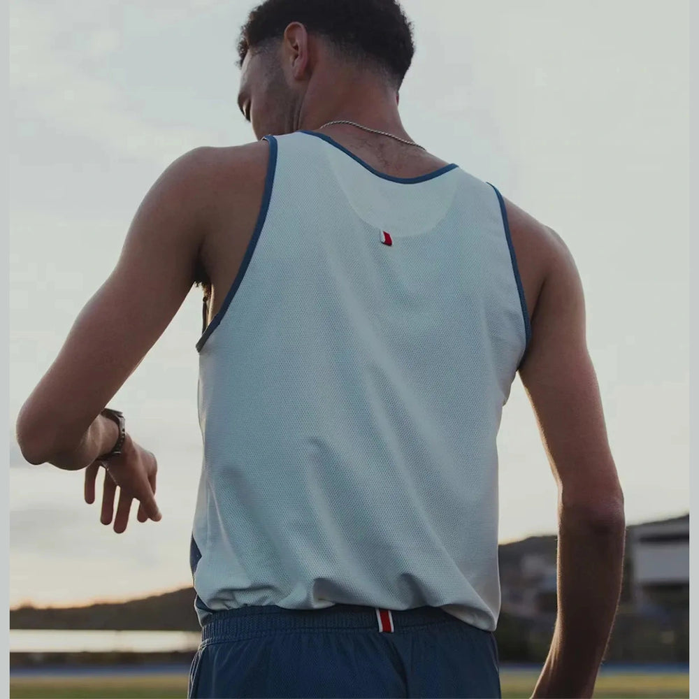 A man viewed from behind wearing a light blue athletic tank top with dark blue trim and a small red logo on the back, standing outdoors on a field at sunset.