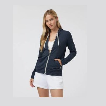 A woman modeling a navy blue Vuori Halo Perform Hoodie 2.0 – Women's with white drawstrings and a white front zipper, worn over a white sports bra and white athletic shorts, against a light gray studio background.