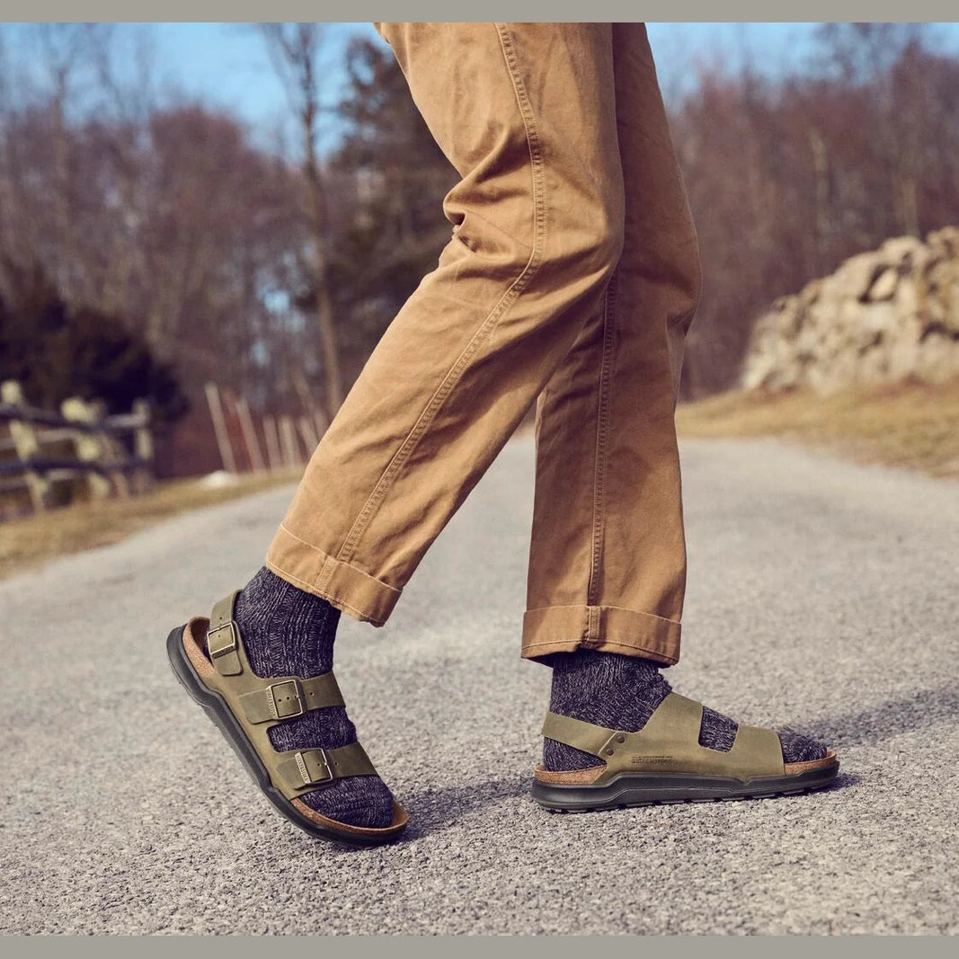 A person walking outdoors on a gravel path wearing tan pants, dark socks, and olive green slide sandals with dark soles.