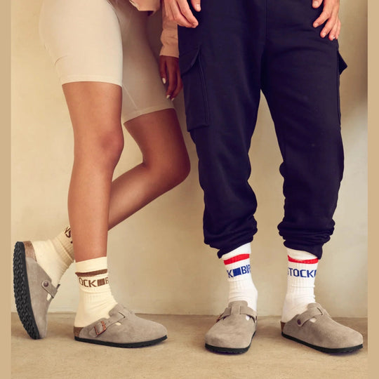 Two people standing indoors wearing white socks with blue and red stripes, paired with gray slip-on clogs. One person also wears beige biker shorts, and the other wears dark navy jogger pants.