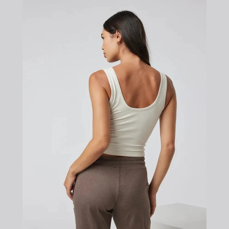 A woman shown from the back wearing a fitted sleeveless Vuori Halo Essential Tank – Women's tank top in a light cream color and brown pants, set against a clean light gray studio background.