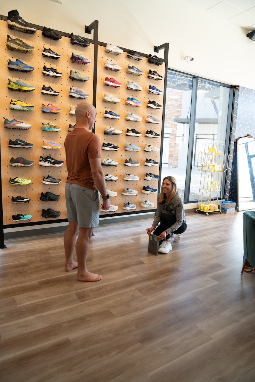 A man in a brown t-shirt and gray shorts stands barefoot on a wooden floor, looking at a woman kneeling down to examine a shoe inside a shoe store. A large wall display filled with various colorful athletic shoes is visible behind them.