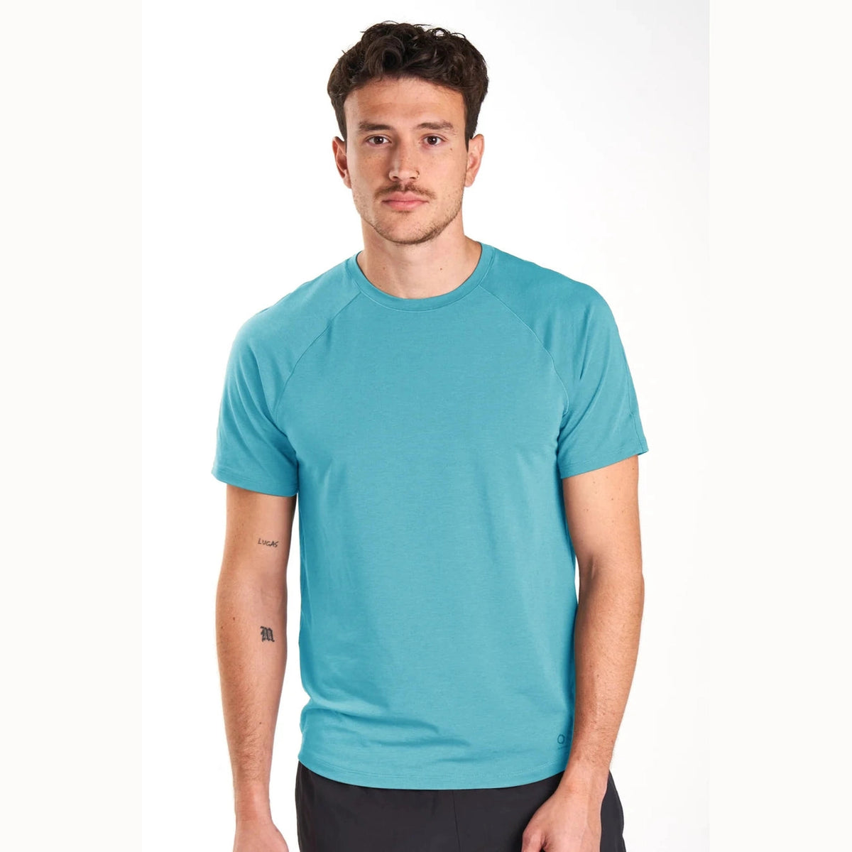 A man wearing a short-sleeve Alwrld Alrn Raglan Tee – Men's in a vibrant turquoise blue color, paired with dark gray shorts, photographed against a clean white studio background.