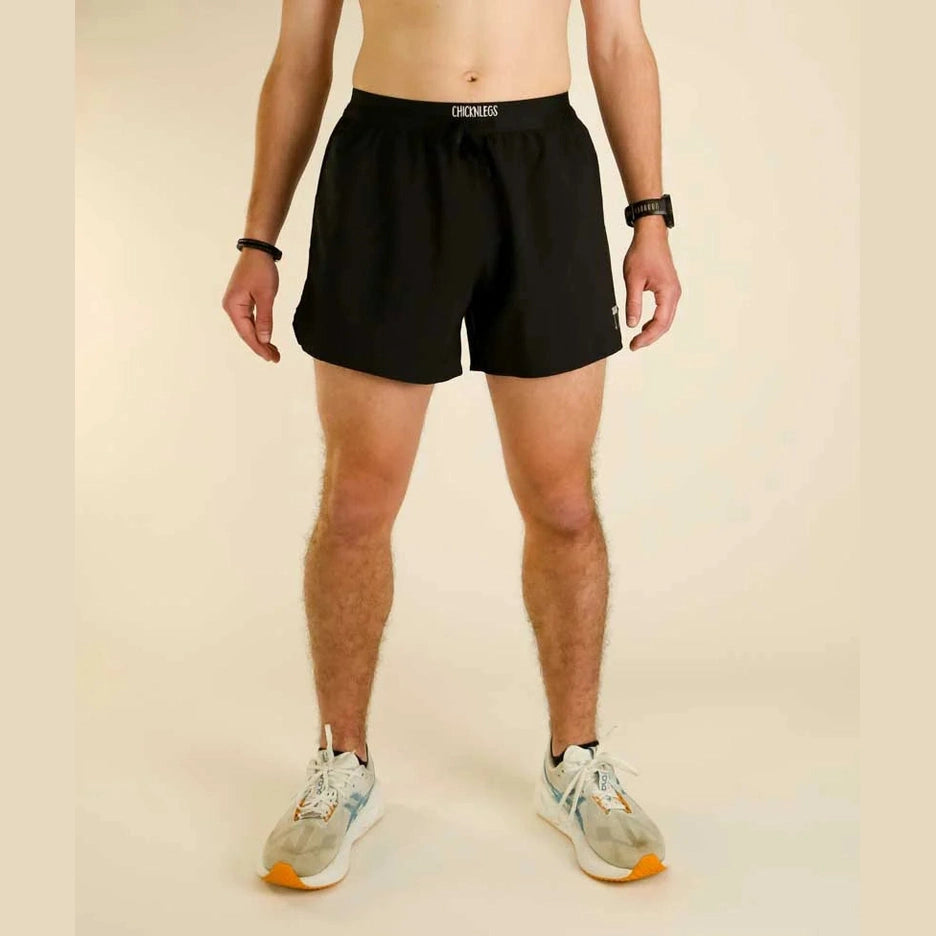 A man wearing black Chicknlegs 5" Running Shorts – Men's with a white 'Chicknlegs' logo on the waistband, paired with gray running shoes, standing against a solid beige background.