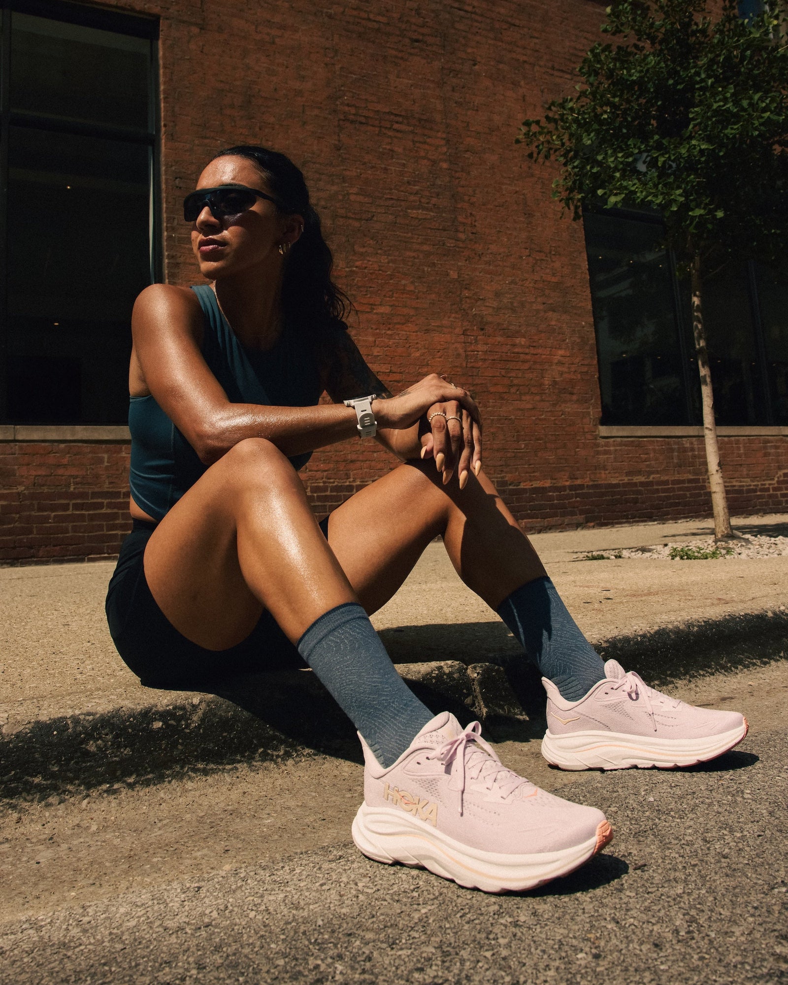 Woman sitting on a sidewalk wearing athletic attire and pink  Hoka Women's Clifton 10 sneakers in front of a brick building.