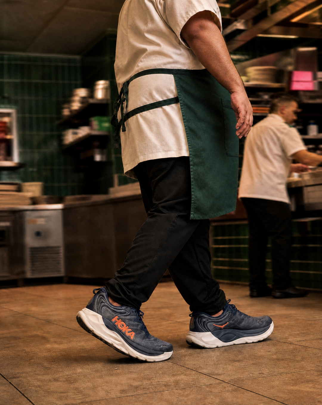 Person wearing a green apron and blue sneakers with Hoka Mens Gaviota 6 in a kitchen setting.