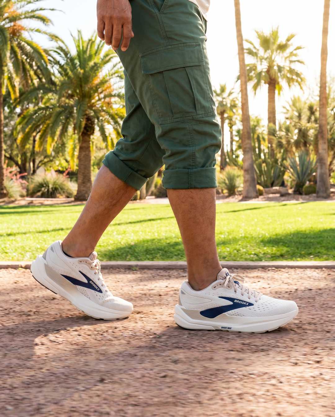 Person wearing green cargo shorts and Brooks Ghost Max 3 white sneakers with blue accents, walking on a path with palm trees in the background.