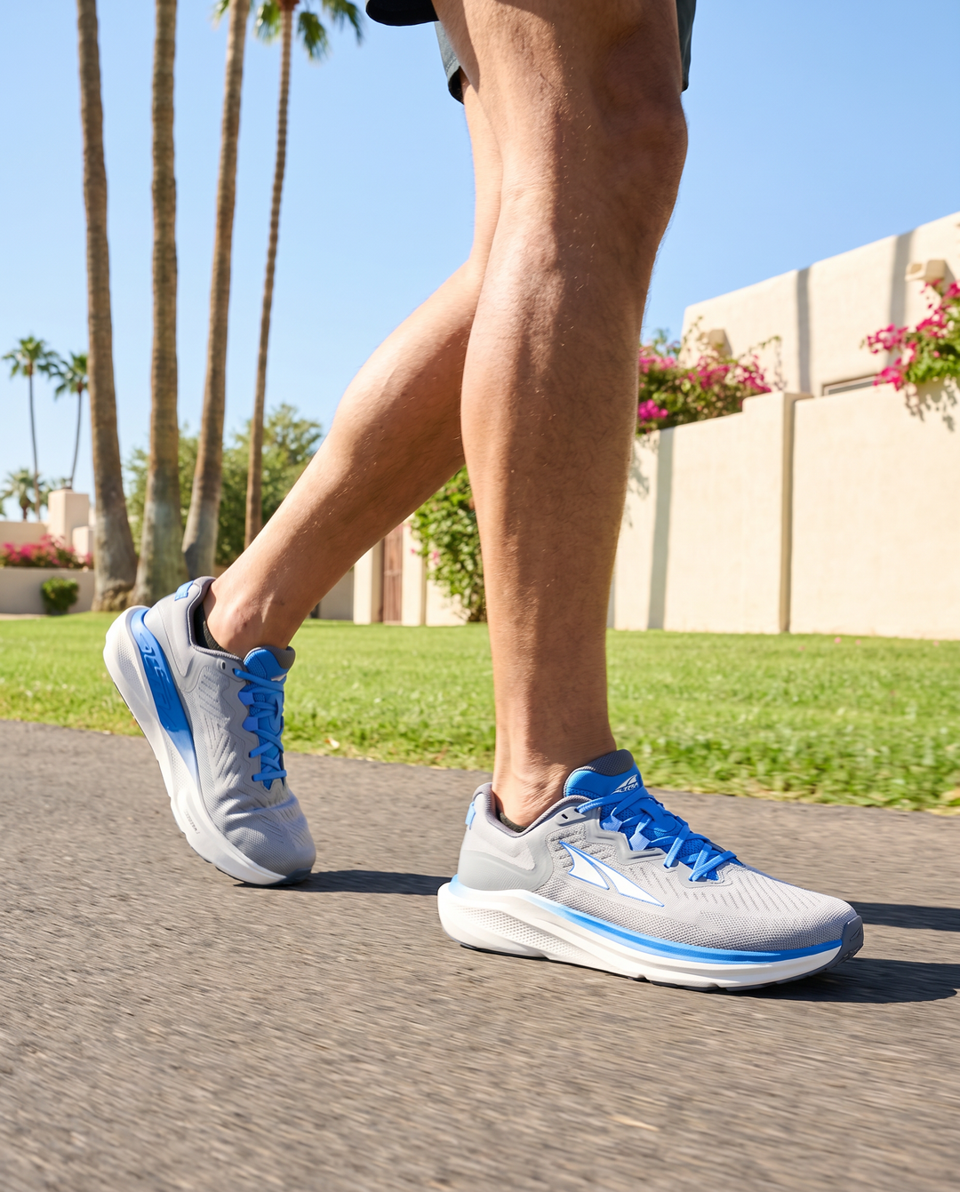 Person wearing gray and blue running shoes on a paved path with palm trees and a building in the background.