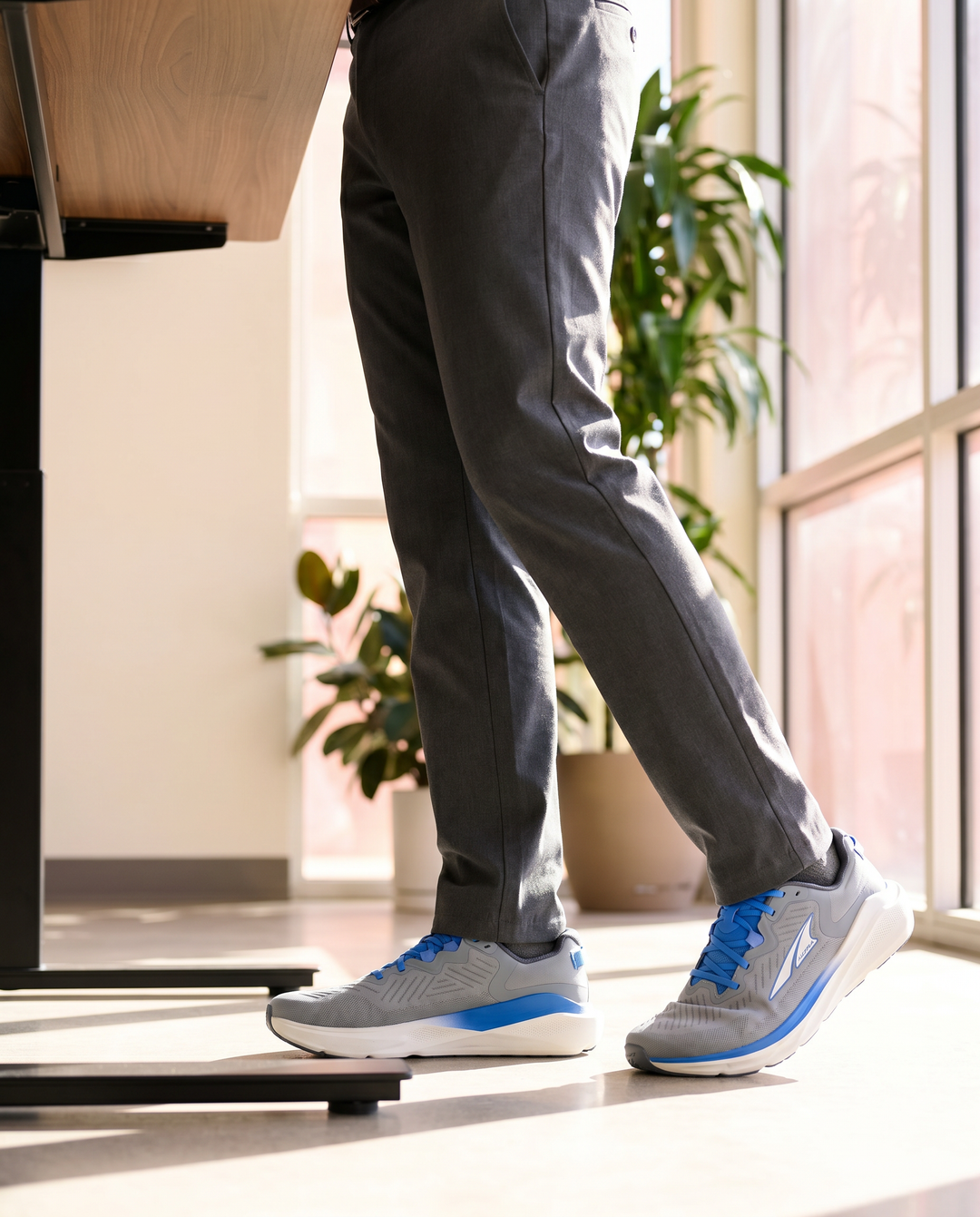 Person wearing gray pants and blue sneakers standing indoors with a plant in the background