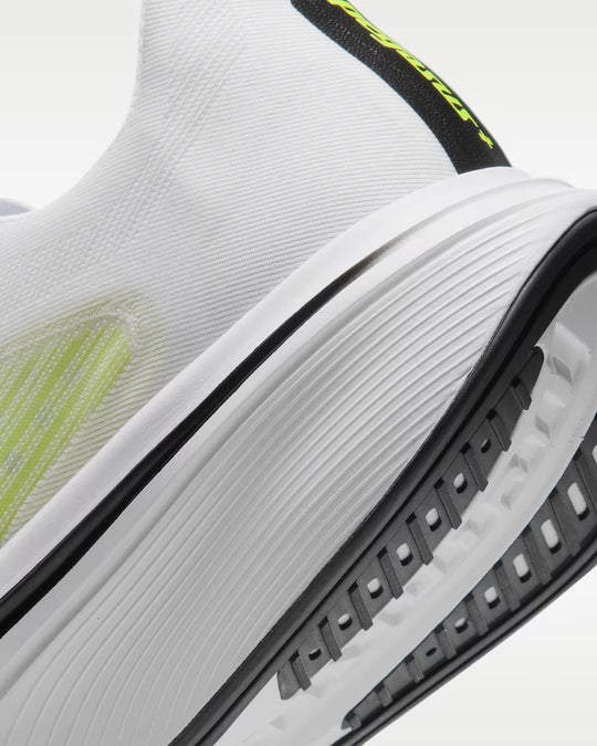 Close-up of a white athletic shoe showing the textured knit upper, thick white midsole, black outsole, and neon yellow-green accents.