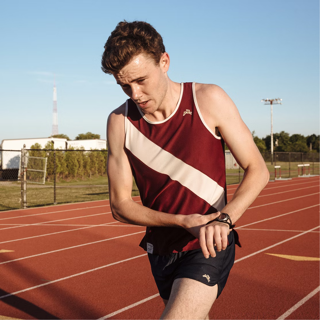 A male athlete on an outdoor running track wearing a sleeveless athletic tank top in maroon and cream, paired with dark shorts. He is looking down at his wrist watch, with the red running track and blue sky in the background.