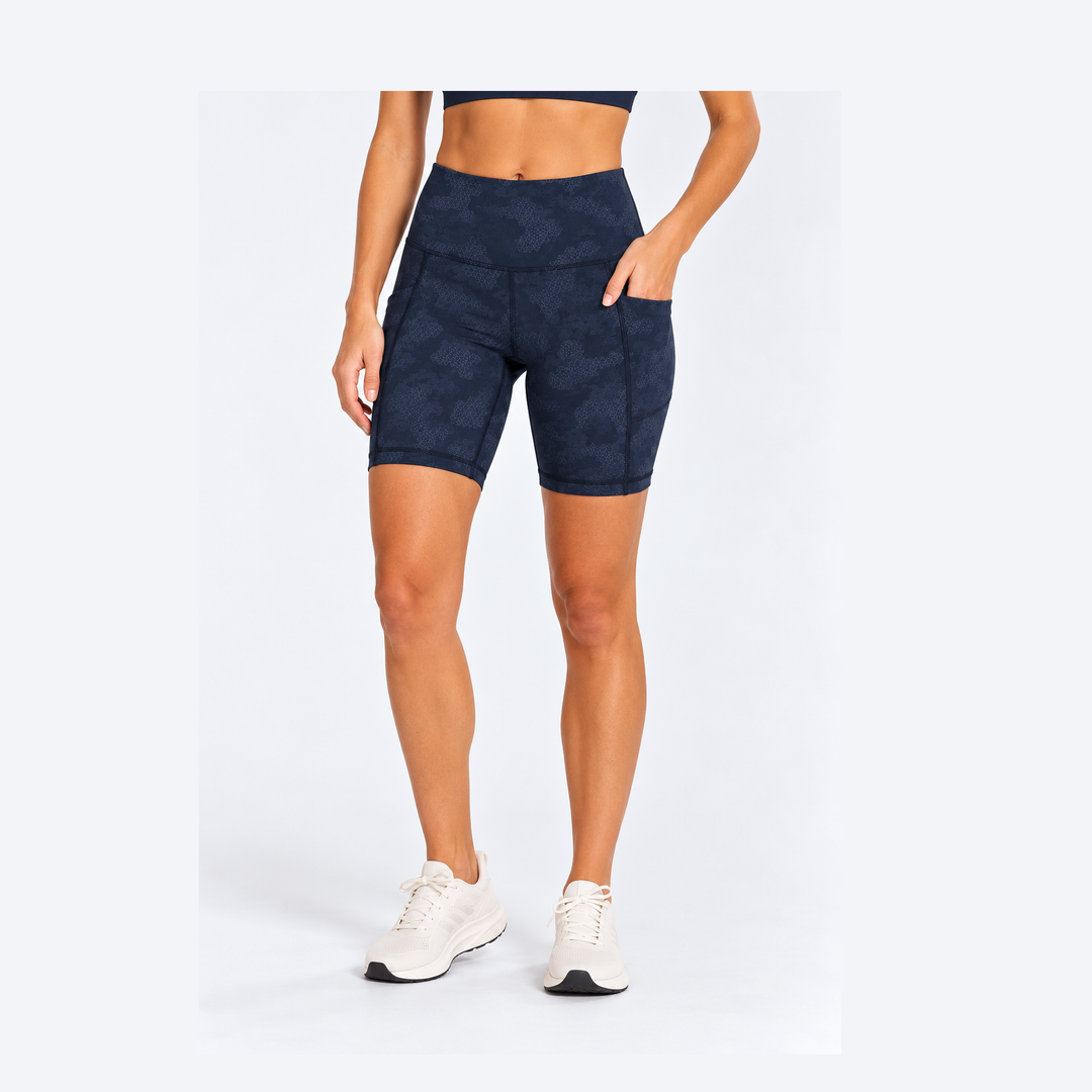 A model wearing 7" Running Bike Short – Women's patterned biker shorts with side pockets, paired with white athletic sneakers, shown from the waist down on a light gray studio background.