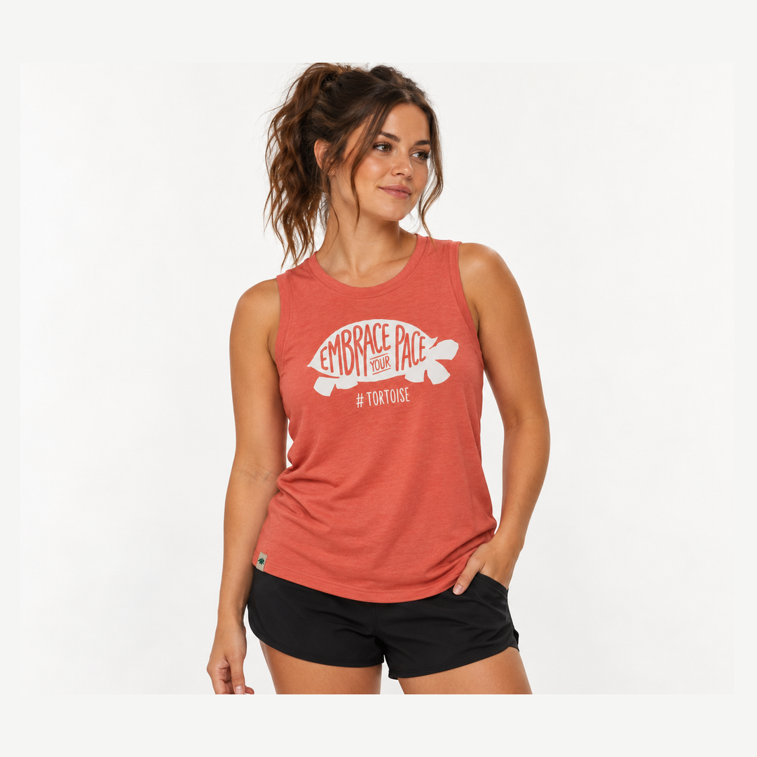 A woman wearing a heathered tank top with a white turtle graphic and the text 'EMBRACE PACE' on the front, paired with black athletic shorts, against a plain white studio background.