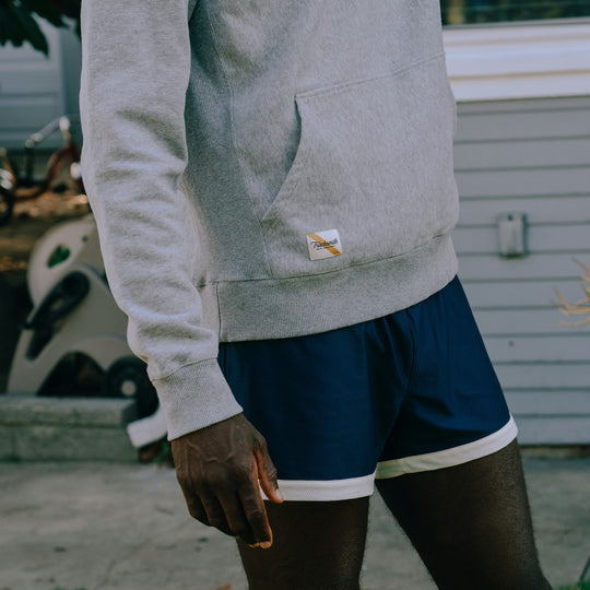 A person wearing a light gray pullover hoodie and navy athletic shorts with a white stripe, standing outdoors in front of a light gray house wall.