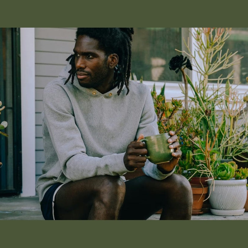 A man with dreadlocks sits on a porch wearing a light gray long-sleeve henley shirt and holding a green ceramic mug, surrounded by potted plants.