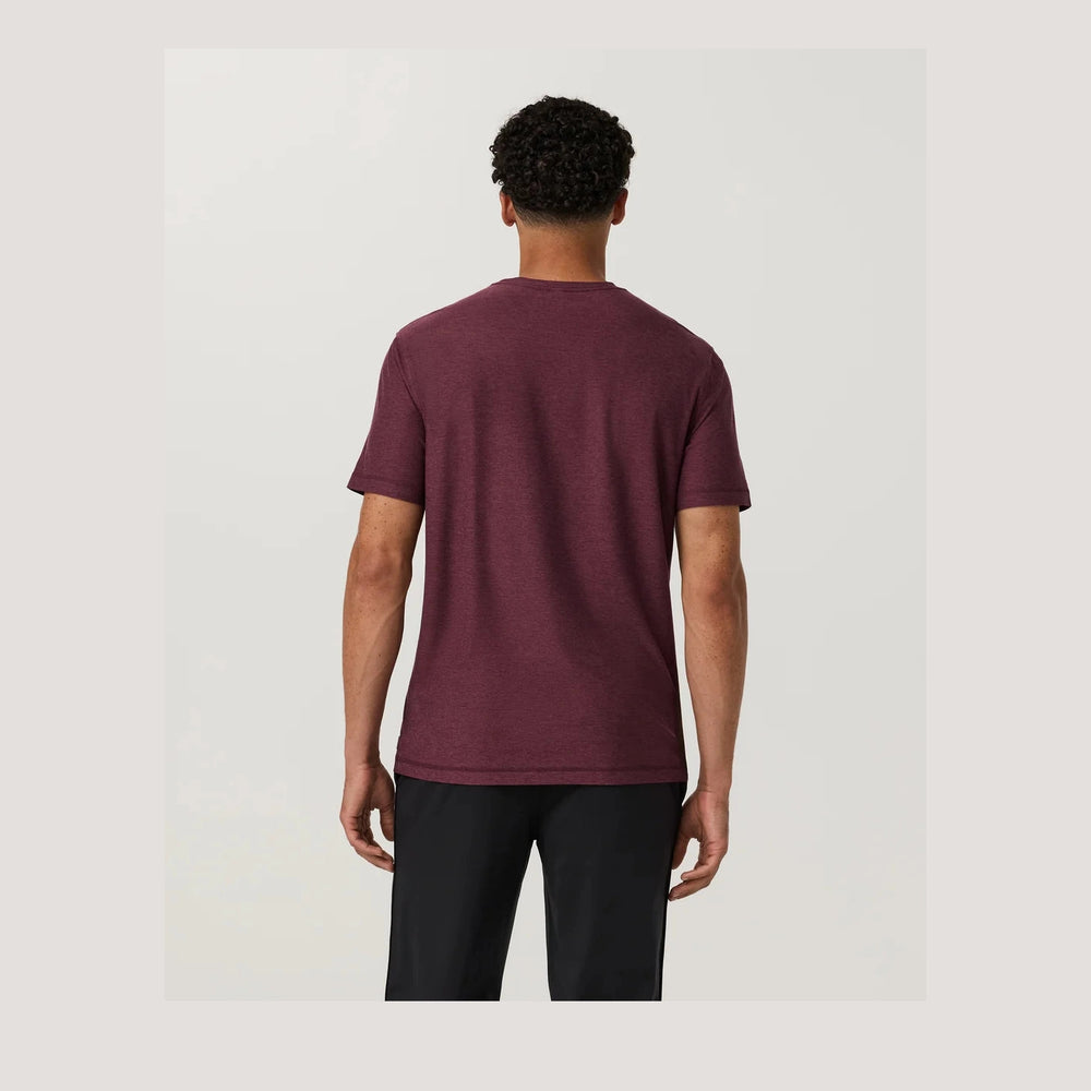 A man viewed from behind wearing a Strato Tech Tee – Men's in heathered burgundy and black pants, standing against a light gray studio background.