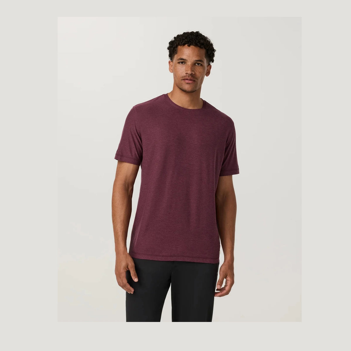 A man wearing a Strato Tech Tee – Men's in heathered burgundy, paired with black pants, standing against a light gray studio background.