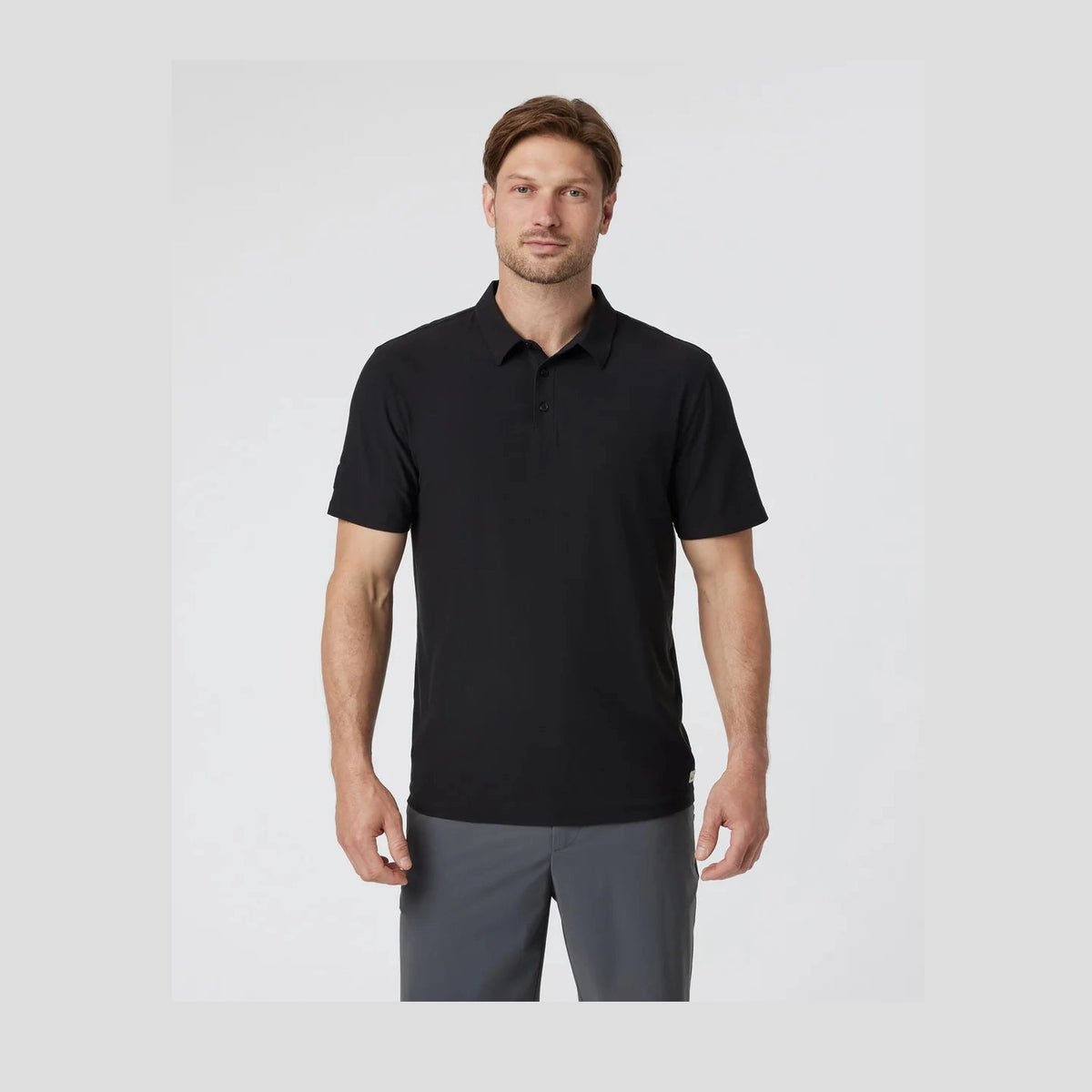 Man wearing a black polo Vuori Strato Tech Polo – Men's shirt and gray pants on a light gray background