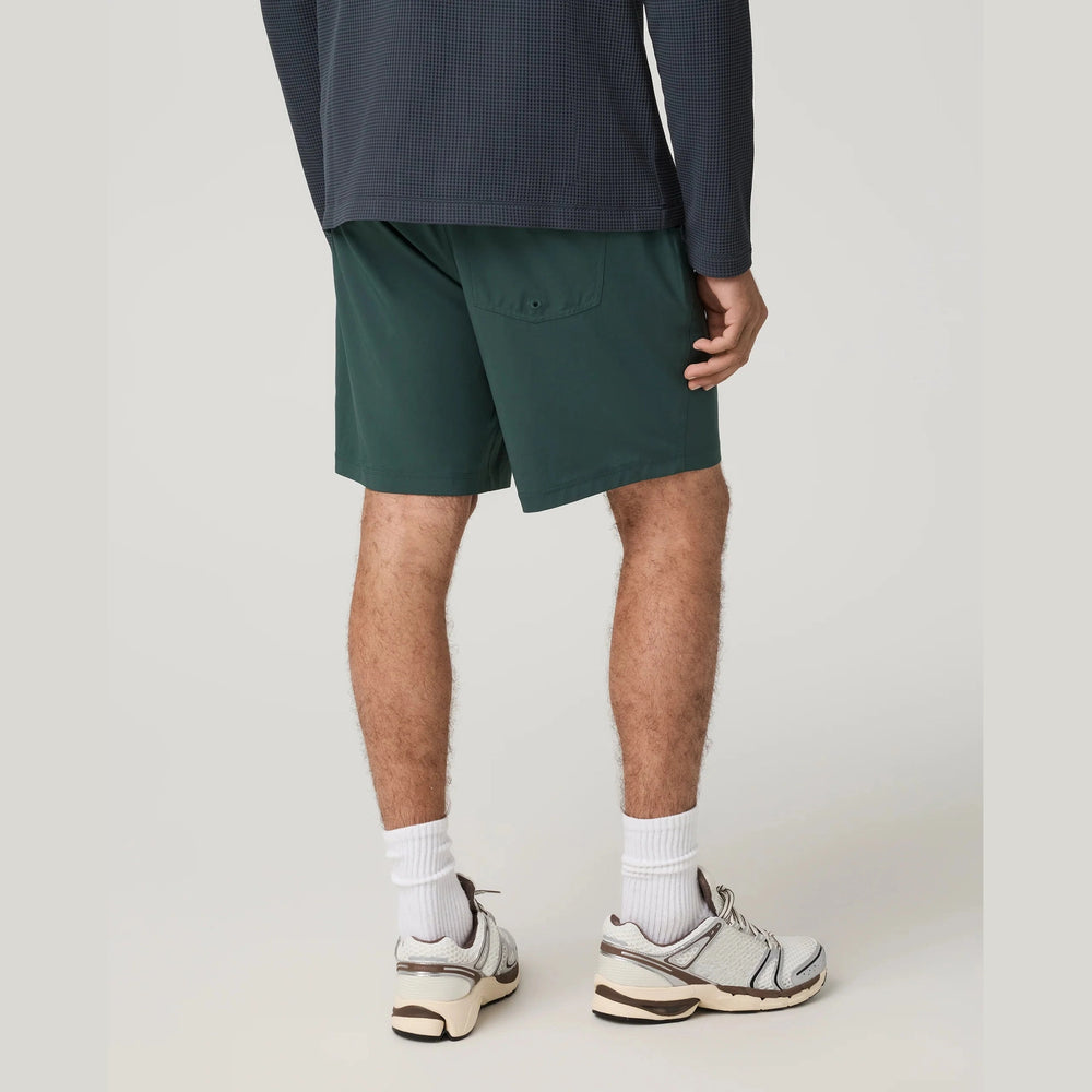A person shown from the waist down wearing dark green Kore Short 7.5" – Men's athletic shorts, a dark navy long-sleeve top, white socks, and white and gray athletic sneakers against a light gray studio background.