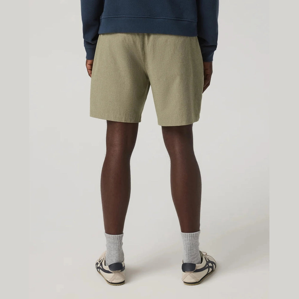 A person shown from the back wearing light olive green Vuori Aim Elastic Waist Short – Men's shorts, a navy sweatshirt, gray socks, and white sneakers with navy accents, photographed against a plain light gray studio background.