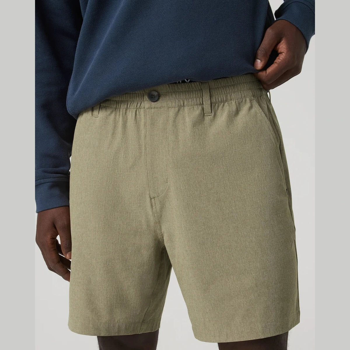 A close-up of a person wearing olive green Vuori Aim Elastic Waist Short – Men's athletic shorts and a navy long-sleeve top, cropped from the chest down against a light gray studio background.