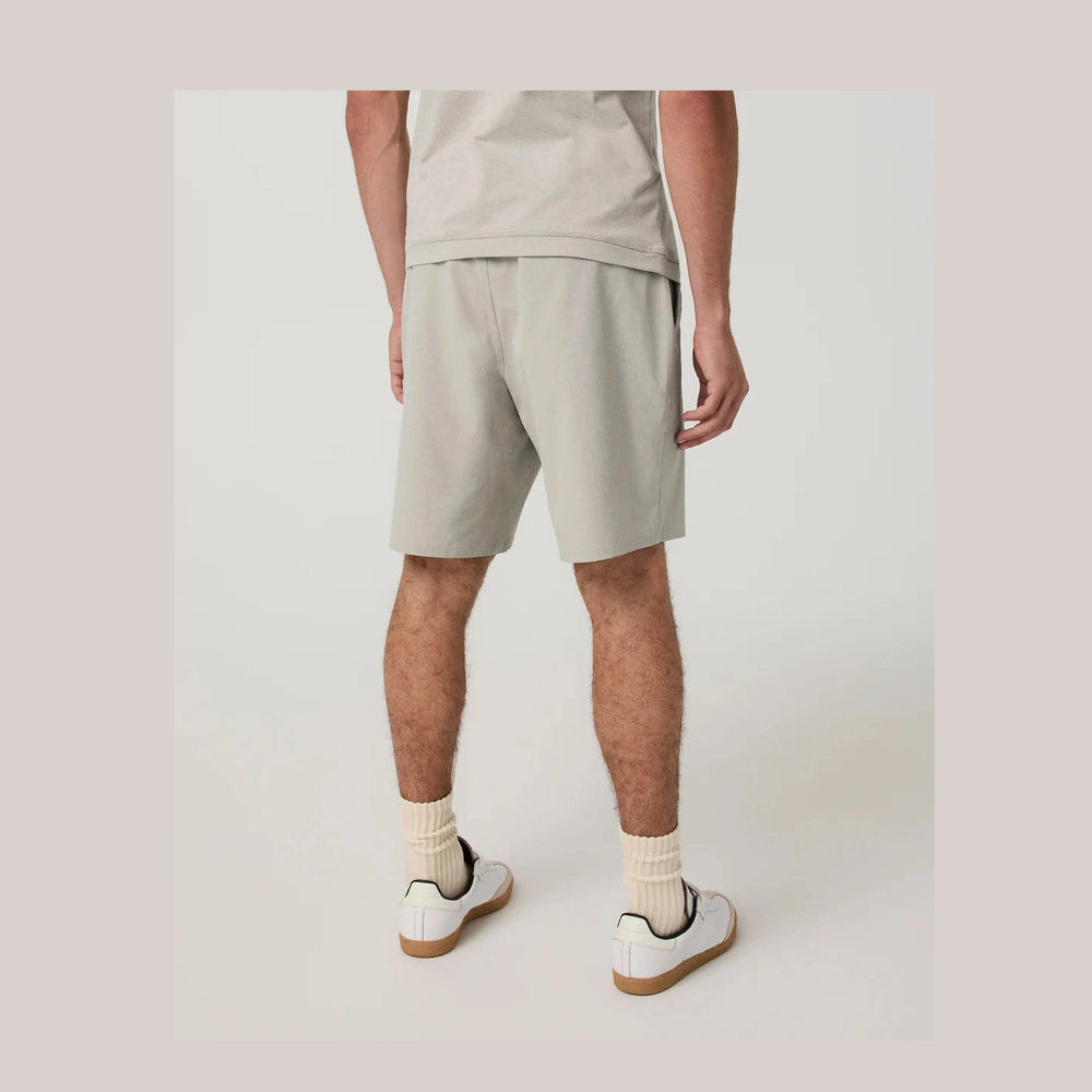 A person shown from the back wearing light beige Vuori Aim Short 8" – Men's athletic shorts, a matching light beige top, cream socks, and white sneakers with gum soles, photographed against a plain light studio background.