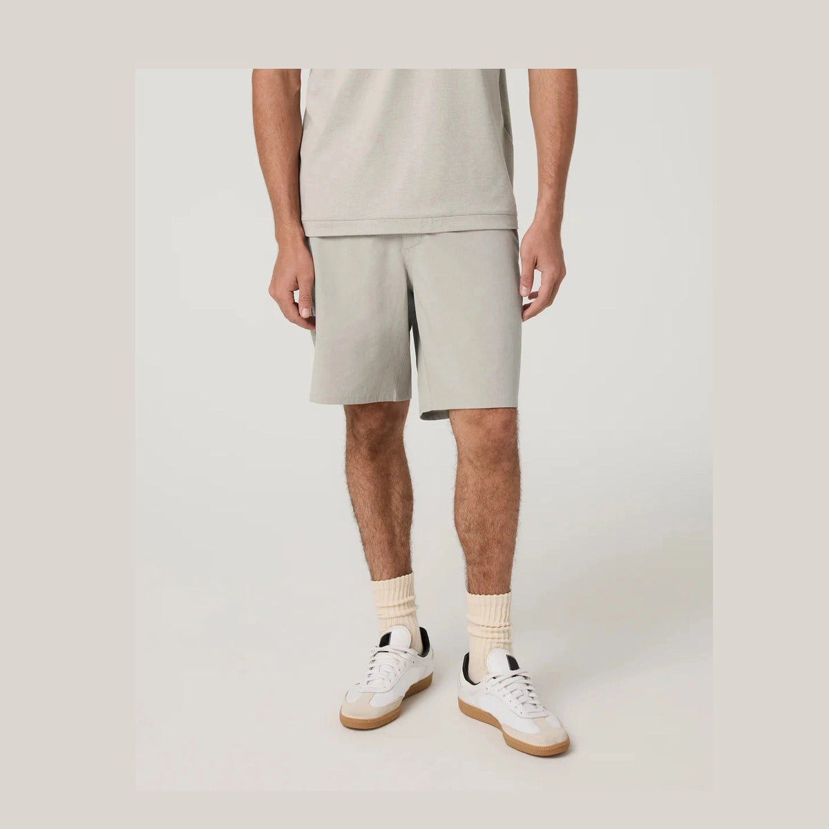 A model from the waist down wearing light beige Vuori Aim Short 8" – Men's athletic shorts, a matching light beige t-shirt, cream ribbed socks, and white low-top sneakers with gum soles, set against a solid light gray studio background.