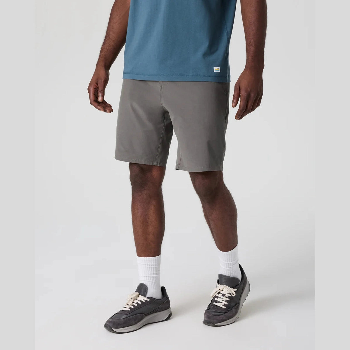 A person wearing gray Vuori Aim Short 8" – Men's shorts, a blue t-shirt, white socks, and dark gray sneakers, shown from the waist down against a light gray studio background.