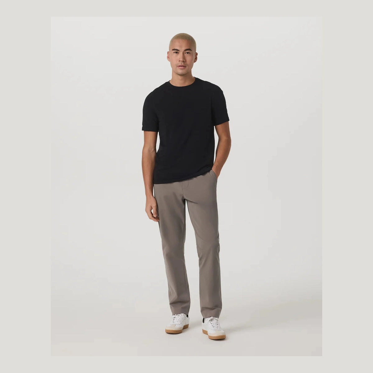 A man modeling a black short-sleeve crewneck t-shirt and light brown Vuori Meta Elastic Waist Pant – Men's trousers with white sneakers, standing against a plain light gray studio background.