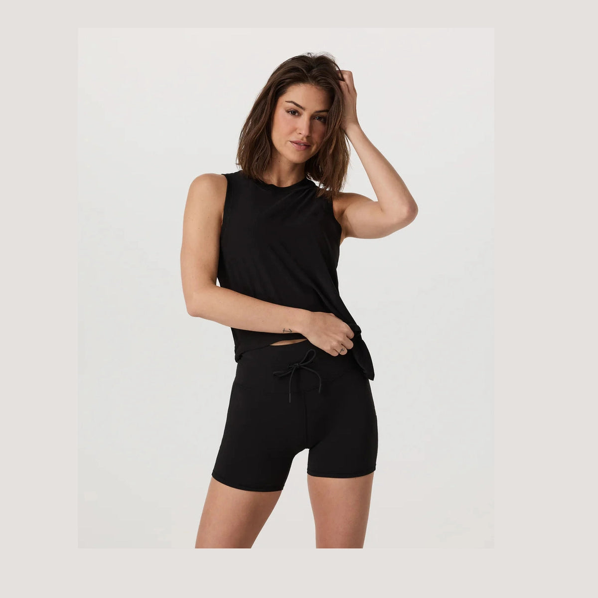 Woman wearing a black Vuori Energy Top Long – Women's sleeveless top and shorts on a light gray background