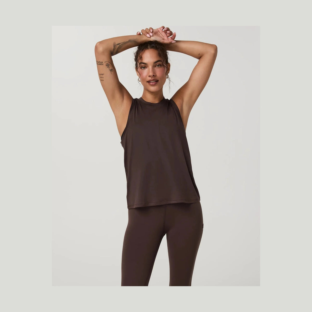 A woman modeling a sleeveless dark brown Vuori Energy Top Long--Women's athletic tank top and matching dark brown leggings against a light gray studio background.