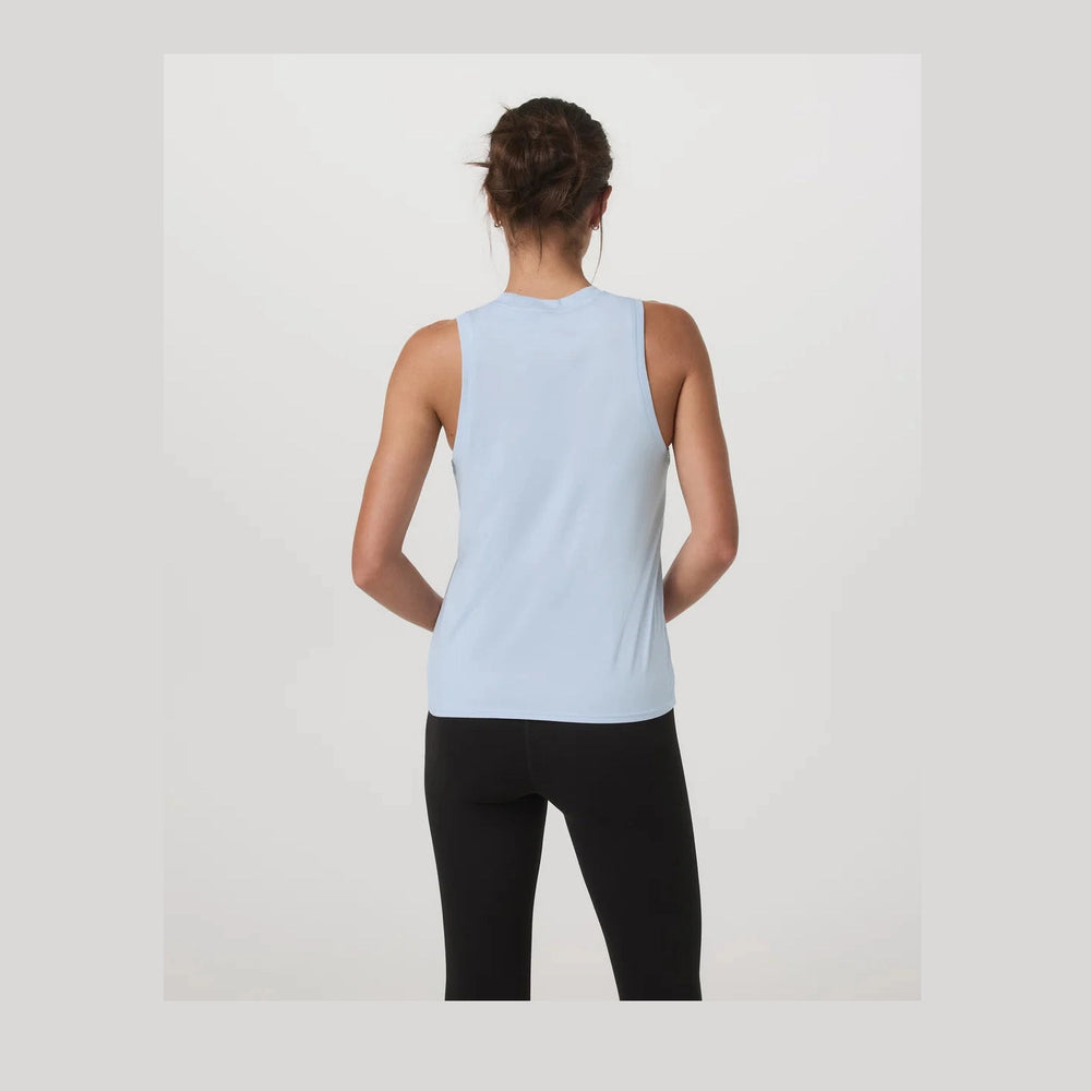 A woman viewed from behind wearing a light blue Vuori Energy Top – Women's sleeveless athletic tank top and black leggings against a plain light gray studio background.