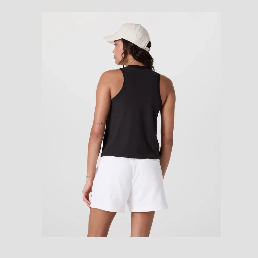 A woman shown from the back wearing a black Vuori Energy Top – Women's sleeveless racerback tank top, white shorts, and a light beige baseball cap against a light gray studio background.