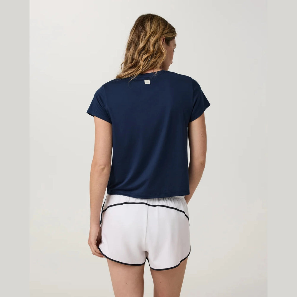 A woman shown from the back wearing a navy blue Vuori Energy Classic Tee--Women's t-shirt and white shorts with black trim, against a plain light gray studio background.