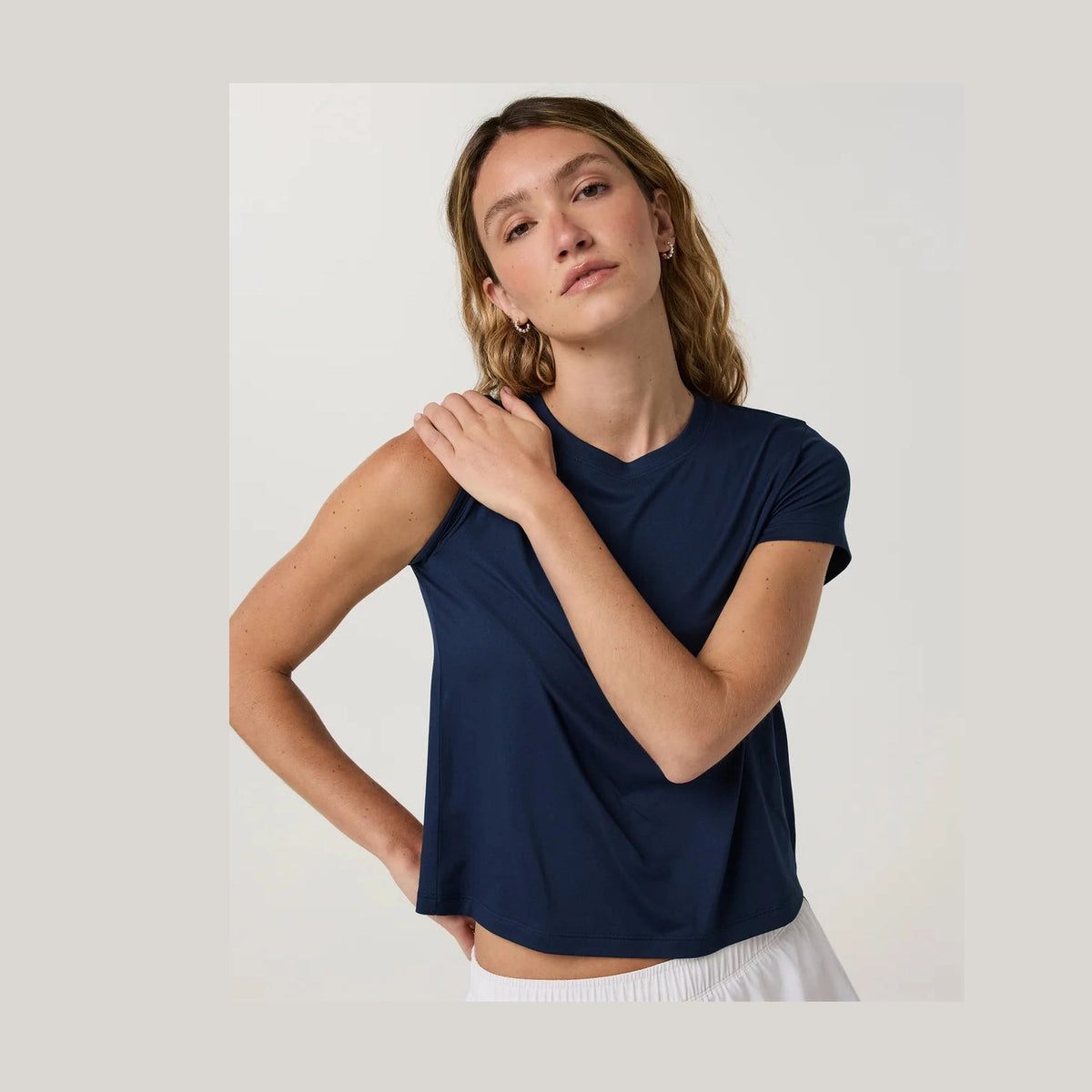 A woman wearing a navy blue Vuori Energy Classic Tee--Women's athletic top, posing with one hand on her shoulder against a plain light gray studio background.
