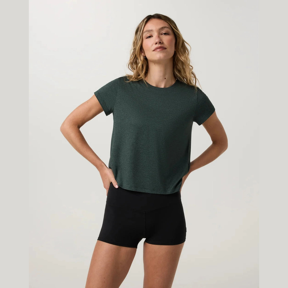 A woman modeling a dark green Energy Classic Tee--Women's athletic top paired with black shorts, standing against a light gray studio background.