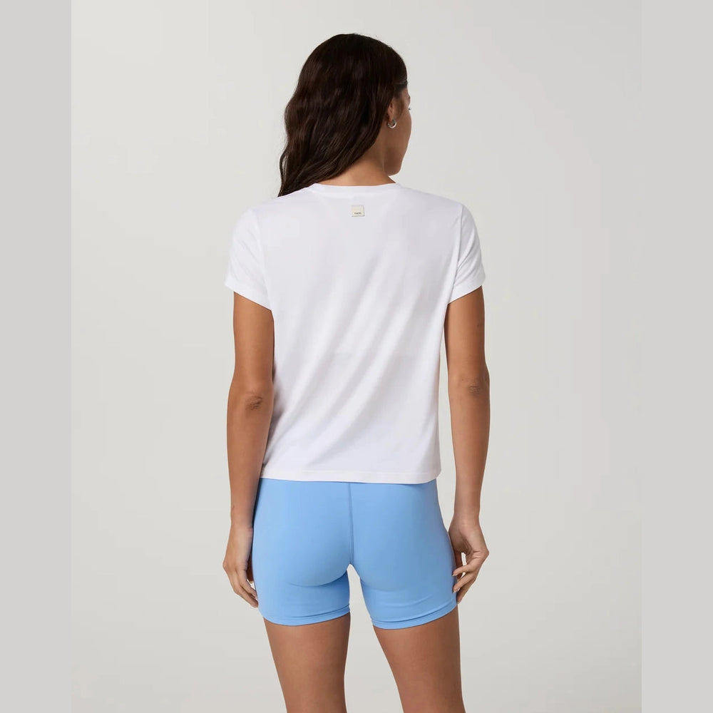 A woman shown from the back wearing a white Vuori Energy Classic Tee--Women's top and light blue fitted shorts against a plain light gray studio background.