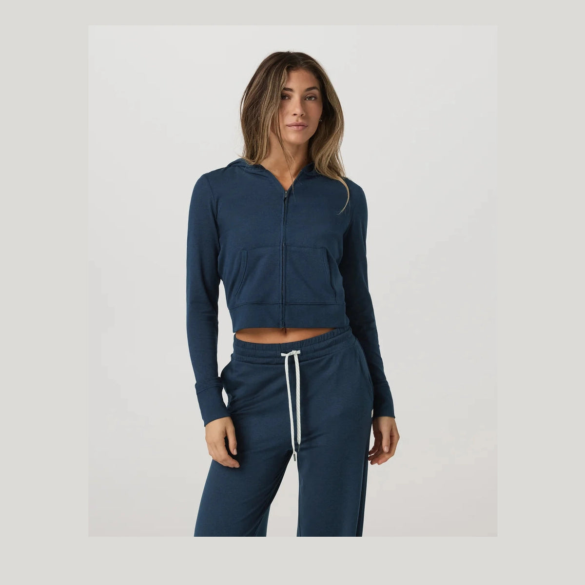 A woman modeling a matching navy blue cropped Vuori Halo Mini Fz Hoodie – Women's hoodie and sweatpants set with a white drawstring, shot against a light gray studio background.