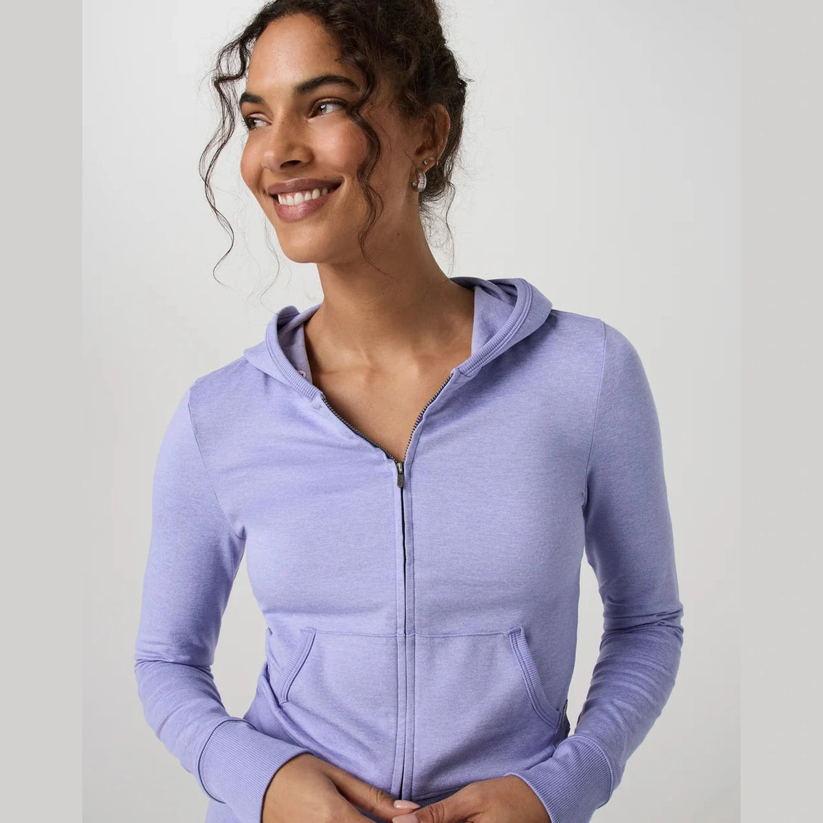 A woman wearing a light purple heather Vuori Halo Mini Fz Hoodie – Women's hoodie, smiling and looking to the side against a plain light gray studio background.