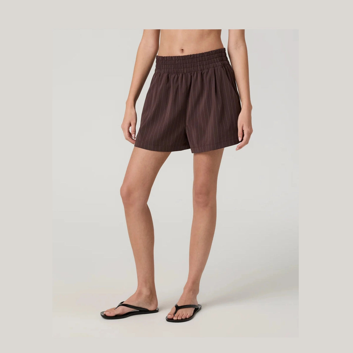 A model wearing dark brown Vuori Villa Short--Women's pinstriped shorts and black flip-flops against a light gray studio background.