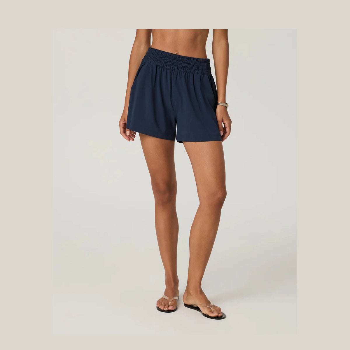 A model wearing a pair of navy blue high-waisted Vuori Villa Short--Women's shorts with an elastic waistband, paired with flat sandals, shown from the waist down against a light gray studio background.