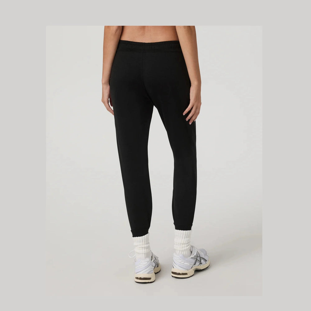 Rear view of a model wearing black Vuori Performance Jogger – Women's jogger pants, white socks, and white sneakers against a light gray studio background.