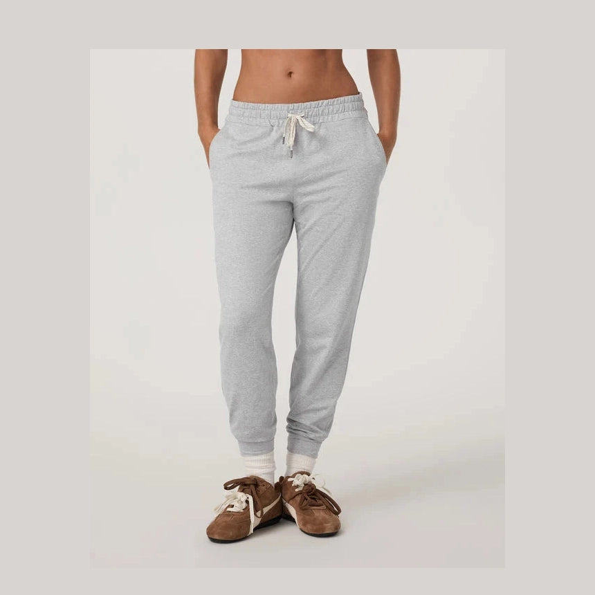 A person wearing light heather gray Vuori Performance Jogger – Women's jogger sweatpants with a white drawstring, paired with white socks and brown slip-on shoes, shown from the waist down against a plain light gray studio background.