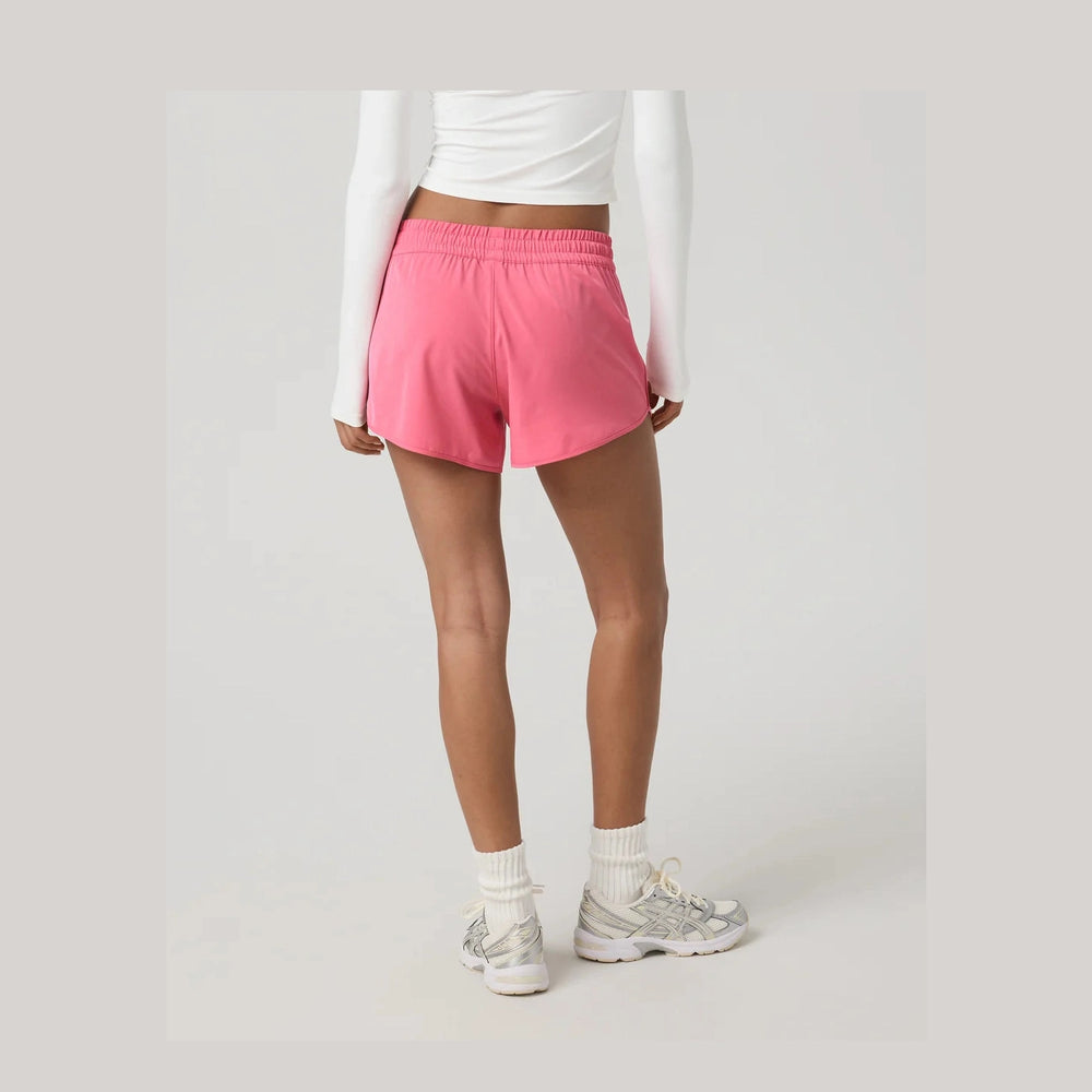 A model from the back wearing bright pink Vuori Clementine 4" Short – Women's athletic shorts, a white long-sleeve crop top, white socks, and white sneakers, set against a plain light gray studio background.